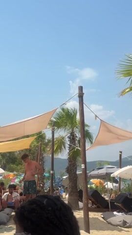 A video clip shows a beach scene with several people visible. In the foreground, a person's braided hair is visible. Further back, two young men in swimwear are on the sand. One is standing and looking towards the left, while the other is walking away from the camera towards the right. Palm trees and beach umbrellas provide shade. The sky is clear and blue with some clouds. The overall setting appears to be a resort or beach club.