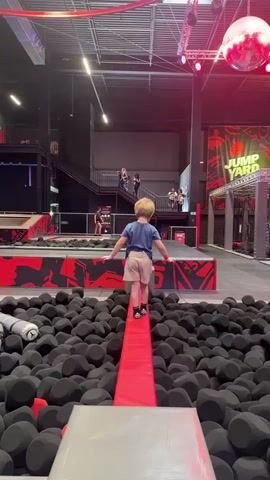 In a brightly lit indoor trampoline park, a young boy walks along a narrow red padded beam surrounded by black foam blocks. He wears a blue shirt, tan shorts, and black socks with a skeleton foot pattern. He carefully places one foot in front of the other, arms outstretched for balance, stepping up onto two gray rectangular blocks along the way.  A girl with blonde hair in a ponytail sits in the foreground, watching the boy. In the background, other people can be seen on an elevated walkway and stairs, some looking down at the trampoline area. A large disco ball hangs from the ceiling, and the words "JUMP YARD" are visible on a sign.