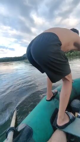The video begins with a close-up of a person in black shorts and flip-flops leaning over the side of a green inflatable boat. The camera then pans to show the boat moving through water, creating splashes. In the distance, a line of green, tree-covered mountains is visible under a cloudy sky. The camera then focuses on a person swimming in the water ahead of the boat. The boat continues to move forward, and the swimmer is visible in the distance.