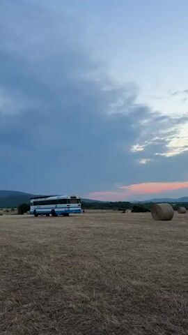 The video opens with a wide shot of a dry, harvested field under a cloudy sky. A blue and white bus is parked in the distance. The camera then pans across the field, showing several round hay bales scattered throughout. As the camera continues to move, the landscape shifts to reveal rolling hills covered in greenery, followed by a view of the sea with rocky outcrops. The sky transitions through shades of blue and pink, suggesting either dawn or dusk. There are no people visible in the video.