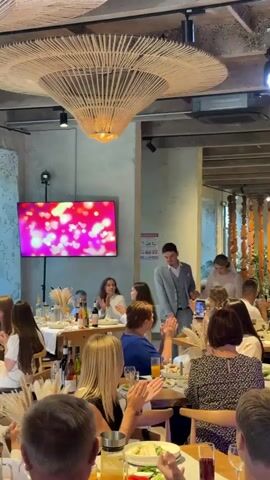 A wedding reception is underway in a well-lit indoor venue. A couple, the bride and groom, enter from the right side of the frame, walking towards the left. The groom is wearing a light grey suit, and the bride is in a white wedding dress and veil. They are walking past tables filled with guests who are seated and appear to be clapping and watching them. In the background, a large screen displays a pink and red heart pattern. The ceiling features a distinctive, large, woven light fixture. The tables are set with plates, glasses, and decorative grasses. The atmosphere suggests a celebratory event.