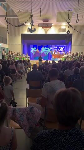 This media file captures a scene from a school play, likely in Ufhusen, Switzerland. The setting is an indoor gymnasium or auditorium, decorated with a pirate theme, featuring hanging anchors and chains. A stage at the front of the room is set with a backdrop depicting palm trees, a volcano, and a skull and crossbones, suggesting a pirate adventure.

The event appears to be a performance for a seated audience. Many children, dressed in green, are gathered on and around the stage, likely participating in the play. Adults are seated in rows of chairs, watching the performance. The lighting suggests an indoor event, possibly in the late afternoon or evening. The atmosphere seems to be one of community and entertainment, with families and children gathered to enjoy the school's production.

The focus of the image is on the stage performance, with the audience in the foreground and midground. There are no obvious weather cues as it is an indoor event. The emotions of the audience are not clearly discernible from this image, but the presence of children and the staged event suggests a positive and celebratory occasion.

Visible text includes "UNTER" written on a backdrop behind the performers on stage, and what appears to be "UFHVSEN" on a banner on the far left side of the stage.