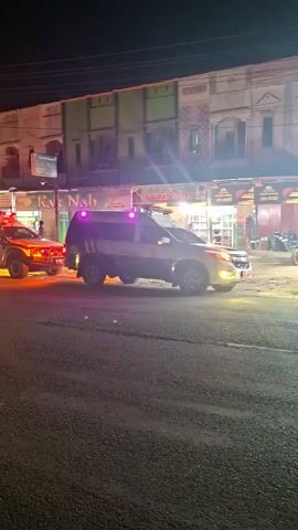 A nighttime scene shows a line of four white utility vehicles and vans on a paved road. The vehicle at the front right has purple flashing lights on its roof and is facing left. Behind it, a white pickup truck or SUV with red and white flashing lights on its roof and front is visible. This vehicle appears to be attached to or towing a third white van with yellow and orange flashing lights. A fourth white van with similar yellow and orange flashing lights is positioned behind the third. The vehicles are stationary or moving very slowly. In the background, multi-story buildings with illuminated storefronts and signs line the street. A person is partially visible near the rightmost vehicle, and another person is faintly visible further down the street on the sidewalk.