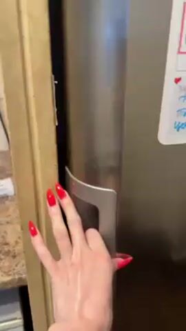 A person with red fingernails opens a refrigerator. The shelves inside are full of food and condiments. There is a mixture of pre-packaged foods, containers, and jars.