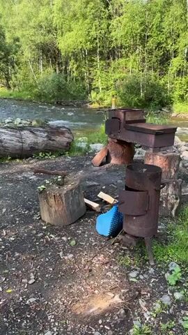 The video opens with a close-up of a rustic outdoor cooking area next to a flowing river. An axe is embedded in a chopping block, with small pieces of firewood scattered nearby. Two rusty metal stoves are present, one larger with a cooking surface and another smaller, cylindrical one. A large fallen log lies horizontally in the foreground, partially obscuring the view of the river. The river flows swiftly through a rocky bed, surrounded by lush green trees and grass. The lighting suggests it is daytime, likely late afternoon due to the warm glow. The overall scene depicts a serene, natural environment set up for outdoor cooking or camping.