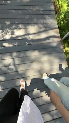 The video shows a first-person perspective of someone walking on a wooden boardwalk. To the left, another person is visible, also walking. The person in the foreground is wearing black leggings and white sandals. The person on the left is wearing blue jeans and white sneakers. The boardwalk is bordered by greenery, suggesting an outdoor, possibly wooded, environment. Sunlight casts shadows on the boardwalk. The camera is moving, indicating walking.