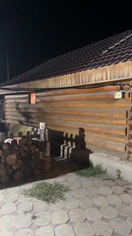 The video pans across a yard at night, illuminated by artificial lights. In the foreground, a paved area with a hexagonal pattern is visible. To the left, a large stack of cut logs is piled against a wooden fence. Behind the logs, a wooden structure with a gabled roof is partially visible. The main focus of the video is a wooden building constructed from large logs, likely a house or cabin. Its wooden exterior is warm-toned and shows the texture of the logs. A small orange window is visible on the side of the building, casting a faint light. To the right of the log building, more stacks of firewood are present. In the background, other buildings and trees are dimly visible against the dark night sky. The lighting creates distinct shadows on the ground and walls of the log building, suggesting a bright light source is positioned to the right.