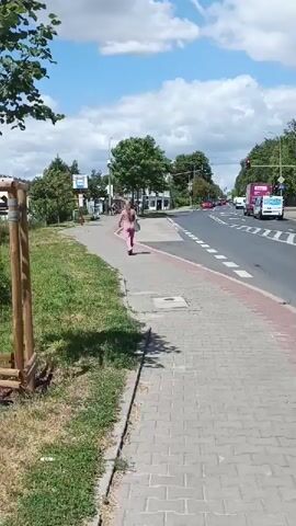 A woman in a pink jumpsuit walks away from the camera on a paved sidewalk. She is carrying a large grey bag over her right shoulder. To her left is a patch of grass and a young tree supported by wooden stakes. To her right is a paved road with a white line marking the edge of the lane. Across the street, there are several small buildings and trees. A blue and white bus stop sign is visible on the left side of the sidewalk. Several electric scooters are parked to the left of the bus stop. A man on a bicycle rides past in the background. The sky is blue with some clouds.