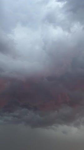 A video shows a dark, stormy sky with thick, gray clouds. A reddish-pink hue is visible through a gap in the clouds, suggesting the sunset or sunrise. The clouds are turbulent and appear to be moving, indicating a change in weather.