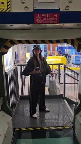 A woman wearing a black hooded jacket, black pants, and a baseball cap stands in a doorway, holding a phone in her left hand and a white shopping bag in her right. She is looking forward. Behind her, the environment appears to be an exhibition or simulation of a spaceship interior, with metallic walls, control panels, and caution signs that read "WATCH YOUR HEAD LOW DOORWAY". The floor is a glass platform showing a representation of Earth below. The camera then pans to the interior of the spaceship, revealing more control panels and equipment. The video continues to pan across the glass floor, showing glimpses of the Earth model and the feet of other individuals standing on the platform.