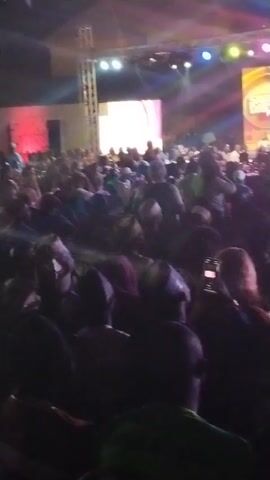 The video shows a large crowd of people gathered in a dimly lit indoor space, likely an auditorium or concert hall. A stage with large screens and lighting is visible in the background, where a figure appears to be performing or speaking. Many individuals in the crowd are holding up their phones, presumably recording the event. The camera moves slightly, offering different perspectives of the crowd and the stage. The lighting is colorful, with beams shining down from the stage.