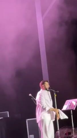 A male singer in a white suit stands on a stage, holding a microphone and what appears to be a piece of paper. He lifts his left arm, waving the paper in the air. A large plume of smoke erupts from below him, illuminated by green stage lighting. The singer is facing away from the camera, looking upwards. A music stand with papers is visible to his right. The background is dark with beams of light.