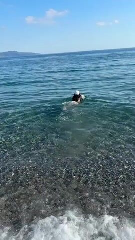 A person wearing a white swim cap and dark swimwear swims in the clear blue-green sea. They are positioned relatively close to a pebble beach, where small waves gently break and recede. The water near the shore is shallow enough to reveal the dark pebbles at the bottom. Further out, the sea deepens to a darker blue. In the distance, a landmass, likely mountains or hills, is visible under a bright blue sky with a few wispy clouds. The scene depicts a sunny day at a beach, possibly in Anatolikos Sinikismos, Greece. No visible text is present.