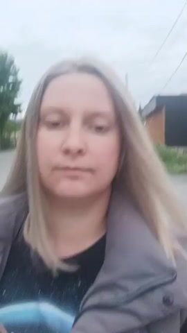 A woman with straight, silver-blonde hair is recording a selfie-style video. She is wearing a black t-shirt with a space-themed graphic and a light gray jacket. She is standing on a street with a small wooden building visible in the background. The sky is overcast. The woman looks directly at the camera and speaks.