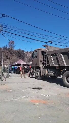The video shows a street with several vehicles passing by. On the right side of the screen is a large brown dump truck. In front of it, there appear to be people standing near a metal fence. Behind them, the side of a hill is visible with some houses. An excavator and two trucks, one gray and one red, also drive by. There are some people cleaning the street. The day is clear and sunny.