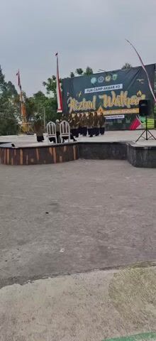 A group of people, likely students, stand on a raised stage in front of a large banner. The banner is black and features text in Indonesian, including "FUN CAMP AKBAR #3" and "HIZBUL WATHAN". Several flags are visible, including the Indonesian flag. Two empty chairs are placed in front of the group on the stage. The area in front of the stage appears to be an outdoor paved area. The sky is overcast.