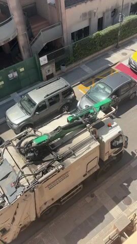 This aerial video shows a garbage truck with a green robotic arm on top parked on a street in Riudoms, Spain. A silver SUV is parked to the left of the garbage truck. A yellow van, possibly a delivery vehicle, is parked in front of the garbage truck. A person on a bicycle is riding in the street behind the garbage truck. The street appears to be paved with concrete and bordered by sidewalks. Buildings with windows and balconies are visible on the right side of the street. Green foliage lines the area in front of the buildings. The weather appears to be sunny and clear, suggesting it is daytime. The scene is likely a residential or commercial area. The garbage truck has "ROS ROCA" written on the side.