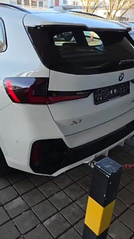 The camera pans from the rear of a white BMW X1 to its side, showing the car parked on a paved lot next to another vehicle. The rear of the car features a BMW logo, X1 badging, and a trailer hitch. The wheels are black with silver accents. The car is parked next to a bollard with a chain. Sunlight creates shadows on the side of the vehicle. The video takes place outdoors during the day.