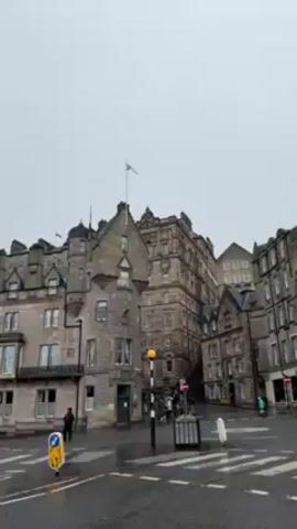 The video shows a street corner in what appears to be a European city, likely Edinburgh. The buildings are constructed of gray stone, with ornate detailing and varying heights, some with turrets or peaked roofs. The sky is overcast and the streets are wet, suggesting it has been raining. People are walking across the crosswalk and along the sidewalks, and a silver car drives by.  There are traffic lights and signs present at the intersection. A small planter is also visible on the corner.