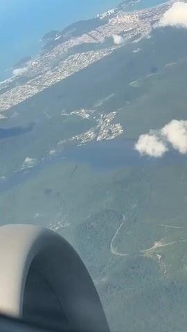 The video shows an aerial view from an airplane window, looking down at a landscape with a coastline, a city, rivers, and dense green forests. The ocean is visible in the distance, a pale blue under a clear sky. A long, narrow city stretches along the coast, characterized by numerous high-rise buildings. Inland, a complex system of dark blue rivers and waterways snakes through vast expanses of lush green forests. Scattered white clouds cast shadows on the landscape below. Part of the airplane's wing or engine is visible in the bottom left corner of the frame. The camera slowly pans upward, revealing more of the distant ocean and the horizon.