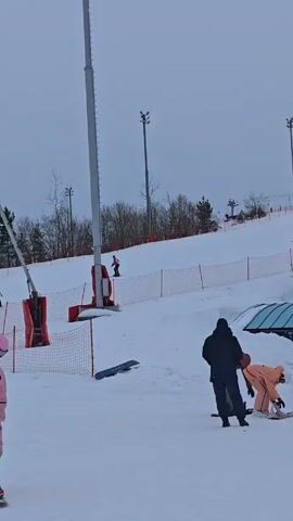 The video shows a ski slope with multiple people skiing and snowboarding. One person, wearing a pink snowsuit and helmet, is snowboarding in the foreground. Other people are skiing or snowboarding further up the slope, and some are walking. There are snow-making machines and safety netting along the slope. The sky is overcast, suggesting it's a winter day.