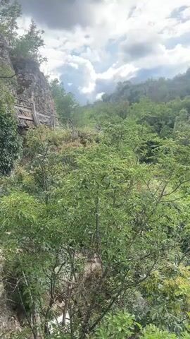 The video pans across a lush green landscape with rocky cliffs and a cloudy sky. In the distance, a waterfall cascades down the rocks, partially obscured by trees. A wooden walkway is visible on the left side of the screen, winding its way up the cliff face. The overall impression is one of natural beauty and rugged terrain.