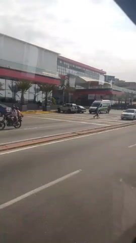 The camera moves forward, passing a large building complex on the left. The building appears to be multi-storied, with a white and red color scheme and glass windows. Palm trees are visible in front of the building. The camera captures a road with multiple lanes and a median, with cars, a van, a pickup truck, and a motorcycle passing in both directions. A person is crossing the road near the van. The sky above is partly cloudy. The video seems to be taken from inside a moving vehicle.
