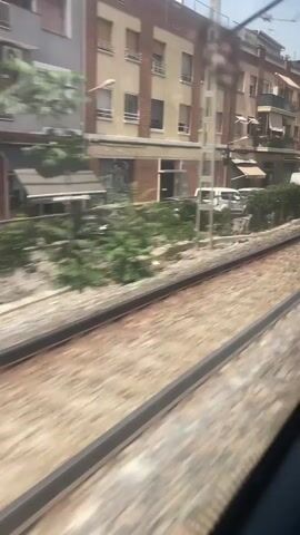 The video shows a view from inside a moving train, looking out a window. The train tracks are visible in the foreground, blurred due to motion. Beyond the tracks, there is a row of buildings with a hedge line separating them from the tracks. The buildings appear to be residential or mixed-use, with windows and balconies. Some white vans and a red car are parked on the street behind the hedge. The overall environment suggests an urban or suburban setting during the daytime. The camera is fixed on the view outside the window, capturing the passing scenery.