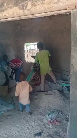 This video depicts scenes from Madugu, Nigeria. The initial segment shows a person in a lime green shirt and light-colored pants operating a machine, possibly for grinding or processing materials, in an indoor setting. The floor is covered with a large pile of what appears to be sand or grain. The room itself is constructed with rough, unplastered walls. The scene is somewhat obscured by dust or haze.

The video then transitions to an exterior view of a more open area. In the distance, a large blue and green truck is visible, suggesting transportation or logistical activity. The surrounding environment appears to be rural, with trees, some buildings, and dirt paths. Power lines are strung between wooden poles, indicating the presence of electricity. The sky is partly cloudy, suggesting it is daytime, likely mid-morning or afternoon. The overall atmosphere seems to be one of everyday work and life in a Nigerian village. No discernible text is visible in the video.