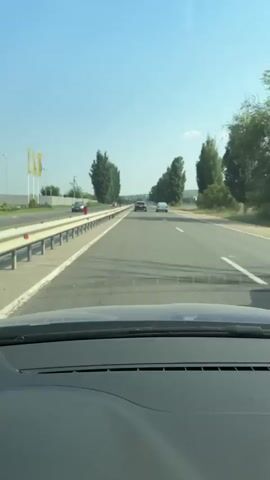 The video shows a first-person perspective from inside a car driving on a highway. The road is paved with clear lane markings and a guardrail on the left side. Tall, slender trees line both sides of the road at intervals, creating a visually rhythmic pattern. Several cars are visible in the distance, moving in the same direction as the camera car. The sky is clear and blue, indicating a sunny day. The dashboard of the car is partially visible in the foreground. The overall scene depicts a journey on an open road under a clear sky.