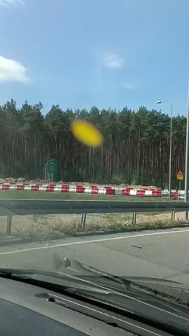 The media file captures a view from inside a vehicle, likely a car, as it travels along a road. The camera is positioned to show the dashboard and windshield, with the outdoor scenery in the background. The scene appears to be a highway or a road with multiple lanes, bordered by a forest of tall pine trees. The weather is sunny with a clear blue sky.

A prominent feature in the middle ground is a row of red and white traffic barriers, suggesting road construction or a temporary lane diversion. There are also streetlights visible, indicating this is likely a public road. A large, blurry yellow object is present in the sky, possibly an insect or a speck of dirt on the windshield. A road sign with a yellow triangle and a black symbol is visible on the right side of the road.

The activity is simply the vehicle in motion, driving past the scenery. The time of day is clearly daytime, given the bright sunlight and clear sky. There are no people or animals visible in the frame. The location cues, such as the type of trees and the road infrastructure, combined with the provided location context of Mała Nieszawka, Poland, suggest an outdoor, possibly rural or suburban, driving environment. No text is clearly legible in the video. The overall mood is neutral, documenting a travel experience.