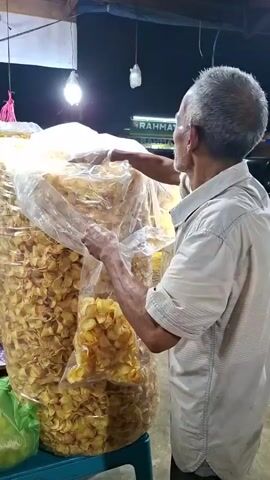 An elderly man with graying hair, wearing a light-colored short-sleeved shirt, is standing at a stall at night. He is holding a large, clear plastic bag filled with golden-brown, flat, circular snacks. He is reaching into another large, clear plastic bag of the same snacks, which is stacked high and behind the first bag. To the left of the man and the snack bags, there is a green plastic bag containing what appears to be a large green fruit or vegetable. In the background, there are more bags of snacks of various colors and shapes, and packets of snacks hanging from the ceiling. There are lights illuminating the stall, and the overall environment appears to be an outdoor market or roadside stall. A woman wearing a headscarf and a light-colored garment walks away from the camera into the background of the scene.