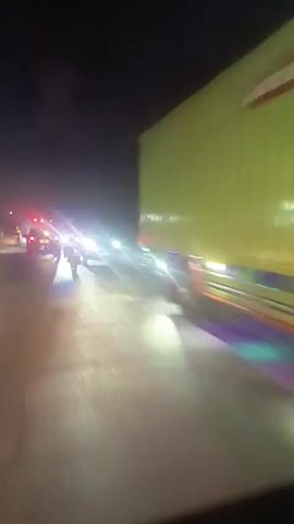 A video taken at night shows a road with several vehicles. The perspective appears to be from inside a car, looking forward. In the distance, there are vehicles with flashing red and blue lights, suggesting emergency vehicles. Headlights from oncoming cars create bright glare. Some vehicles have colorful lights underneath them. The road surface appears wet, reflecting the lights. The sky is dark.