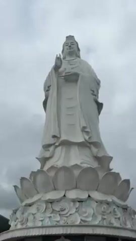 The video shows a very large, white statue of a female figure, presumably a deity, standing on a large lotus base. The sky is overcast. People are walking around the base of the statue, taking photos. Some are ascending the stairs leading to a structure built into the base of the statue. The statue is the focal point of the scene, dwarfing the people and the surrounding environment. The atmosphere is calm and reverent.