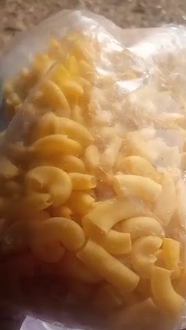 A close-up shot shows a clear plastic bag filled with elbow macaroni. The bag appears to be slightly damp, with condensation visible on the inside. The macaroni is a light yellow color and is packed tightly within the bag. The background is out of focus, but suggests an indoor setting with a countertop and possibly other objects or containers. The lighting is somewhat dim.
