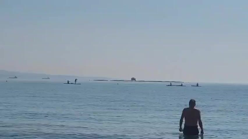 A man stands in the shallow water at the edge of a calm ocean. In the distance, several people are paddleboarding on the water. Further out, there are several large ships on the horizon, and a small island with a building on it. The sky is clear and light blue.