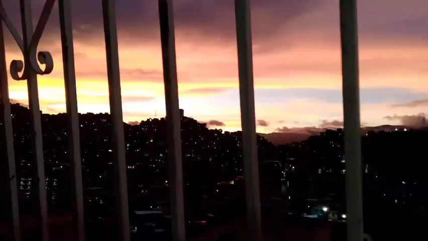 The video shows a sunset over a cityscape seen through a white metal railing. The sky is filled with vibrant colors of orange, pink, and purple, with clouds partially obscuring the sun. Below the sky, a densely populated urban area is visible, with buildings clustered on hillsides and scattered throughout the valley. Many lights are visible in the city, indicating it is dusk or evening. Mountains can be seen in the background, also partially covered by clouds. The railing in the foreground consists of vertical bars and a decorative curved element on the left side. The camera appears to be stationary.