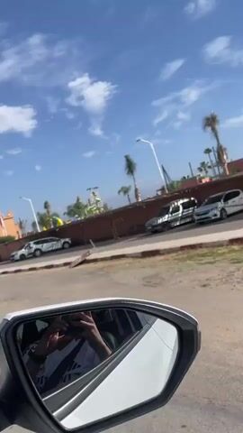 A vehicle drives down a street, passing a water park called "Aqua Fun Club". There are a few parked motorcycles on the side of the road and flags are flying at the entrance to the park. The sky is blue with scattered clouds. The camera is inside a car and filming from the passenger side mirror.