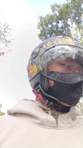 A person wearing a motorcycle helmet and a face mask is riding a motorcycle on a road surrounded by trees. The helmet has a yellow, black, and blue design. The person is wearing glasses and a light-colored hoodie. The road has yellow lane markings. In the background, there are some small structures that appear to be sheds or small buildings. The sky is overcast. The video is shaky, indicating movement.