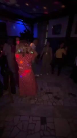 A group of people are dancing in a dimly lit room. The room has a tiled floor and a dark ceiling with exposed beams. Disco lights cast colorful patterns on the floor and walls. In the foreground, a woman in a red floral dress is dancing. Other people, both men and women, are scattered around the room, also dancing or observing. Some are wearing casual clothing. There are framed pictures on the walls and what appears to be a window with curtains in the background. The atmosphere suggests a party or social gathering.