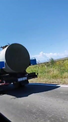 The video opens with a close-up of the rear and right side of an olive green tank truck driving on a paved road. The truck features a large white cylindrical tank on its bed, a blue storage box mounted on the side, and a visible license plate "37 BW 521". The camera, likely from an adjacent vehicle, captures the truck in motion, then pans or overtakes it, showing its full right side. As the truck moves out of frame, the scene transitions to show the roadside, featuring a strip of green vegetation, including tall grasses or reeds, bordering the asphalt. Beyond the roadside vegetation, a gently sloping green and brown hill is visible, extending towards a clear blue sky with a few scattered white clouds. The entire scene is set under bright daylight.