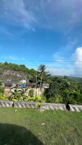 The video shows a landscape view with lush green vegetation and a bright blue sky with scattered clouds. In the foreground, there is a grassy area with concrete barriers and some scattered debris. Several power poles with wires are visible, and some small structures resembling huts or small buildings are nestled into the side of a rocky incline. A palm tree stands prominently on the right side of the frame. The overall scene appears to be a rural or semi-rural setting. The camera appears to be moving, possibly from a vehicle, as evidenced by the slight motion blur and the changing perspective.