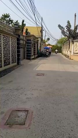 The video shows a residential street lined with houses and fences. Several motorcycles are parked along the side of the road, and two people are standing near a food stall with umbrellas. Another person is riding a motorcycle in the distance. The sky is overcast, and there are many overhead power lines. The road surface appears to be asphalt with some gravel.