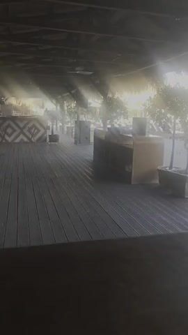 The video shows an outdoor scene at what appears to be a resort or beach bar. The foreground is dominated by a dark wooden surface, possibly a table or bar. Sunlight streams in from the right, creating a bright, hazy atmosphere, with visible light rays. The midground features a wooden deck, with several large, dark boxes or speakers positioned on it. There are also potted trees with sparse foliage scattered around. In the background, there are lounge chairs, umbrellas, and structures that suggest a resort setting. The overall impression is of a tranquil, possibly early morning or late afternoon ambiance. The video is shot from a low angle, looking out towards the open space.