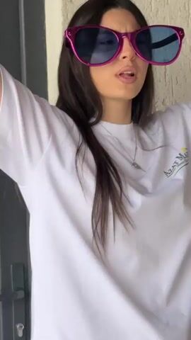 A young woman with long dark hair and large pink sunglasses is shown in a medium shot. She is wearing a white t-shirt with a logo on the left chest. Her right arm is raised above her head, and her left hand is holding a dark rectangular object. Her mouth is open as if she is speaking or singing. The background appears to be an outdoor setting with a textured wall and a dark doorway or column on the left. The lighting suggests it is daytime.