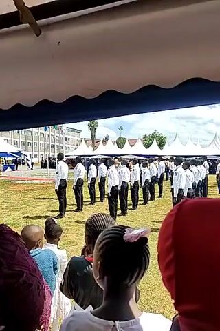 The video shows a formal event taking place outdoors. A line of individuals, dressed in white shirts and black pants, stand in formation on a grassy field. In the foreground, people are seated, presumably as an audience, under a tent-like structure. A tall building is visible in the background. The event appears to be well-organized, possibly a graduation or ceremony.