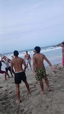 The video shows a group of young men playing volleyball on a sandy beach. They are casually dressed in shorts and some are shirtless. The ocean is visible in the background with waves breaking on the shore. The sky is overcast. The men are hitting the volleyball to each other, with some jumping to reach it. The camera moves, following the ball’s trajectory.