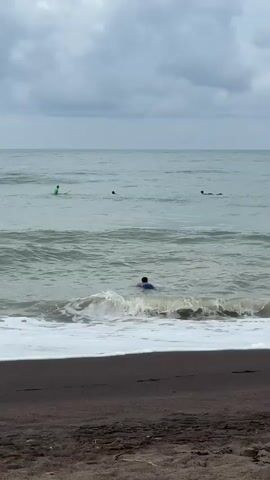 The video shows a beach scene with a person swimming in the ocean. In the background, there are other people in the water, some of whom appear to be on surfboards. The water is choppy with waves breaking near the shore. The sky is overcast. The foreground shows a dark sand beach.