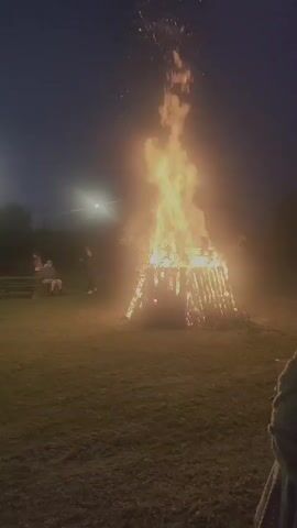 In Lipuški, Latvia, a large bonfire blazes in a grassy field at dusk. The fire is constructed from logs and branches, forming a tall, conical pyre that sends flames soaring into the darkening sky. Sparks and embers ascend from the intense heat, some visible against the twilight.

Several people are gathered around the bonfire, appearing to be watching it. In the background, a group of individuals are seated on what looks like a wooden bench or picnic table. They are silhouetted against the light of the fire, their faces indistinct. The overall atmosphere suggests a communal gathering, possibly a celebration or an outdoor event.

The scene is illuminated primarily by the fire's glow, which casts a warm, orange light across the immediate surroundings. The sky is a deep, dusky blue, indicating that the sun has set and night is falling. A streetlamp or other artificial light source is visible in the distance to the left, providing a small point of contrast to the natural light of the bonfire. The ground is covered in grass, and the silhouette of trees can be seen in the background, suggesting a rural or semi-rural setting.

The mood appears to be one of observation and shared experience, with the focus of attention undeniably on the magnificent, crackling bonfire. The presence of multiple people indicates a social event, where the fire serves as a focal point. The image captures a moment of natural spectacle and human gathering, likely on a cool evening. No visible text is present in the media.