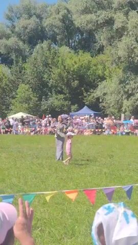 In an outdoor grassy area, an adult wearing a grey suit and a child in a pink dress are standing in front of a crowd of spectators. The adult appears to be holding a cane or a long stick, possibly performing. The child is looking towards the adult. Behind them, tents and more people are visible, indicating an event or festival. Colorful triangular flags are strung across the foreground. The background is filled with lush green trees under a bright sky.
